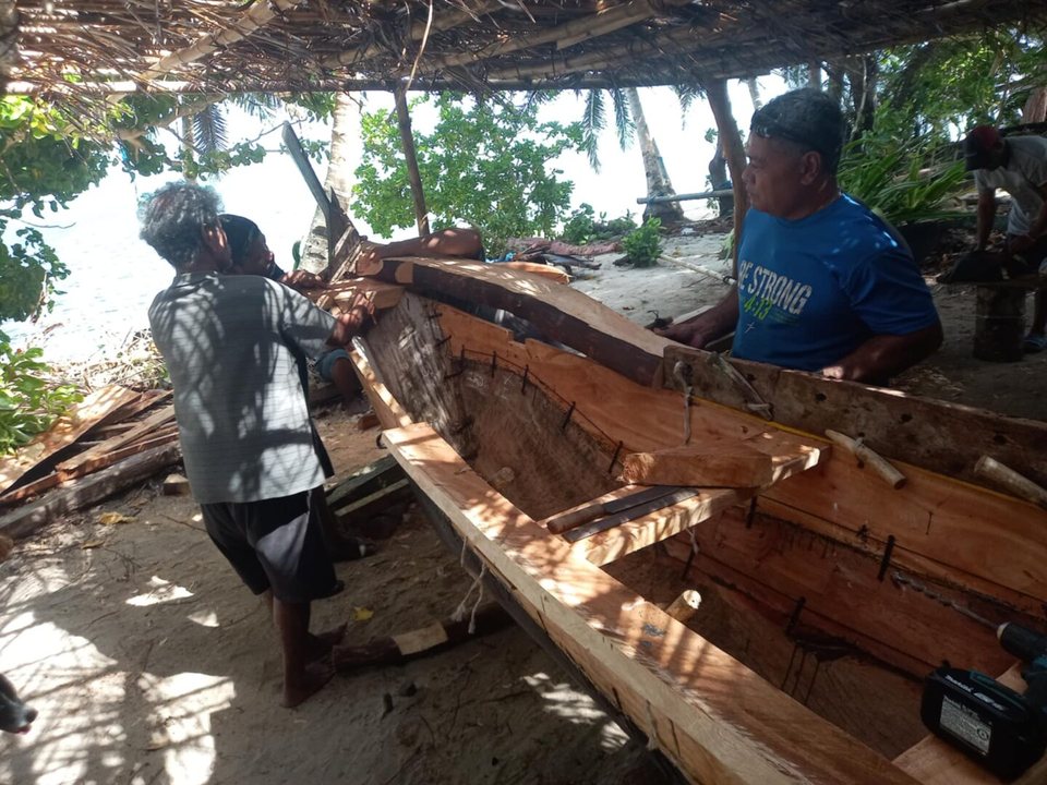 Two men work alongside a wooden canoe hull in a shaded shelter.