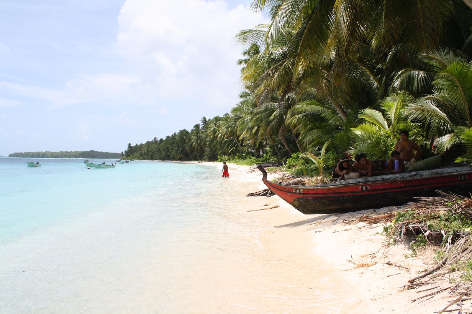 A lagoon shore with coconut palms, small boats, and people gathered beside a red canoe.