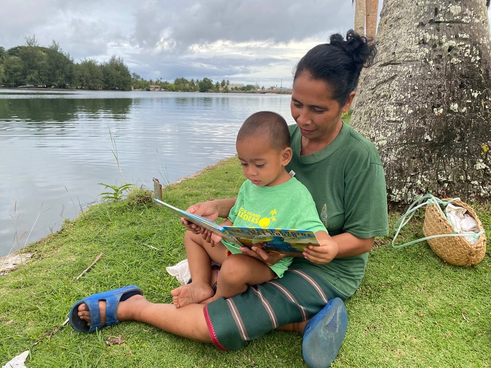 An adult and a young child sit together reading a picture book beside the water.