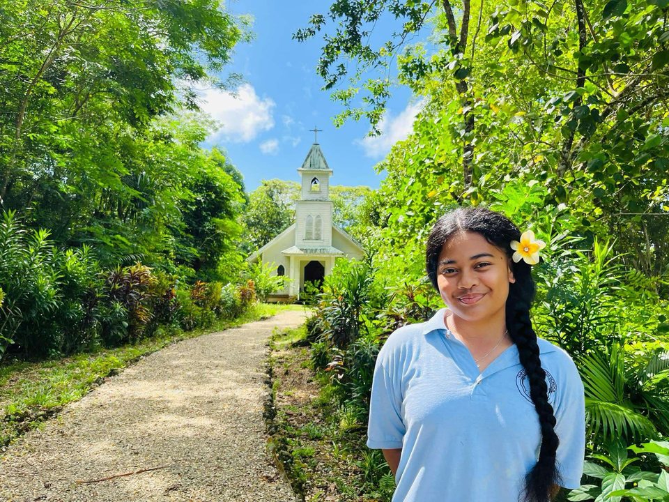 A student in a blue school uniform stands on a leafy path.
