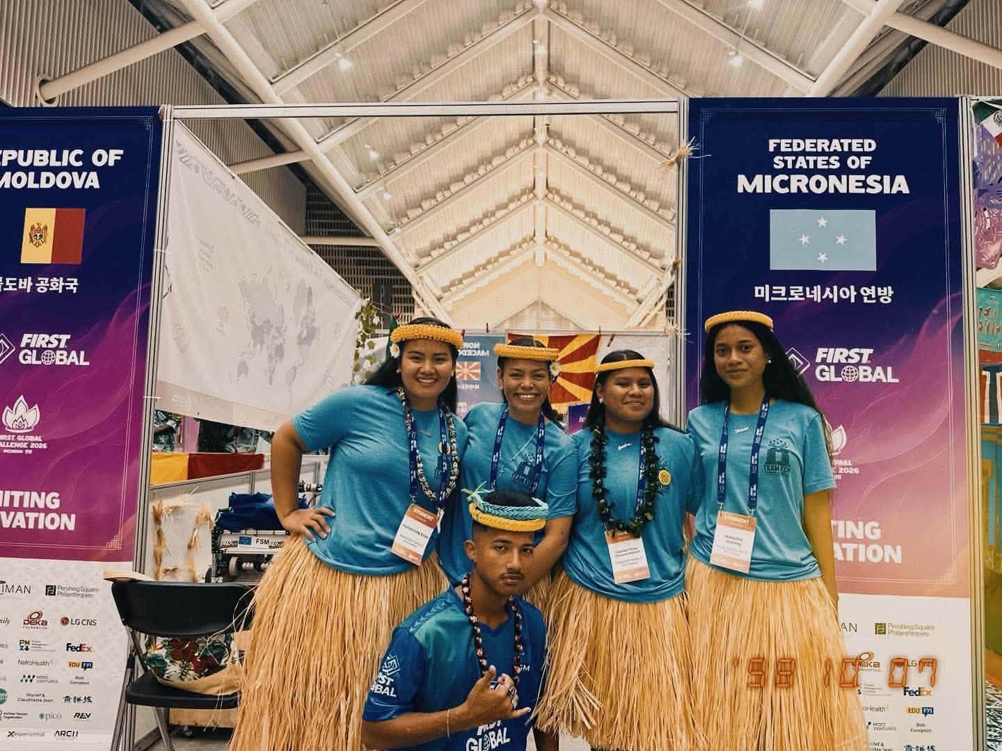 Five Team FSM members in blue shirts and traditional attire pose in front of the Micronesia banner at FIRST Global 2026.